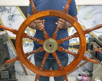 May include: A large, wooden ship's wheel with a brass centre hub and eight spokes. The wheel is brown and has a natural wood finish. The wheel is being held by a person, but the person's hands are not visible in the image.