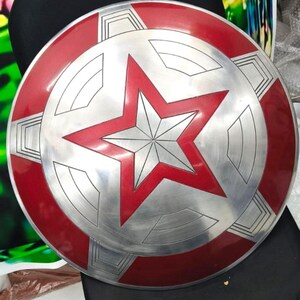 May include: A red and silver Captain America shield with a five-pointed star in the center. The shield has a raised, textured surface.