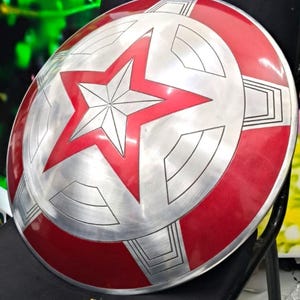May include: A red and silver Captain America shield with a large red star in the center. The shield has a textured surface and is made of metal.