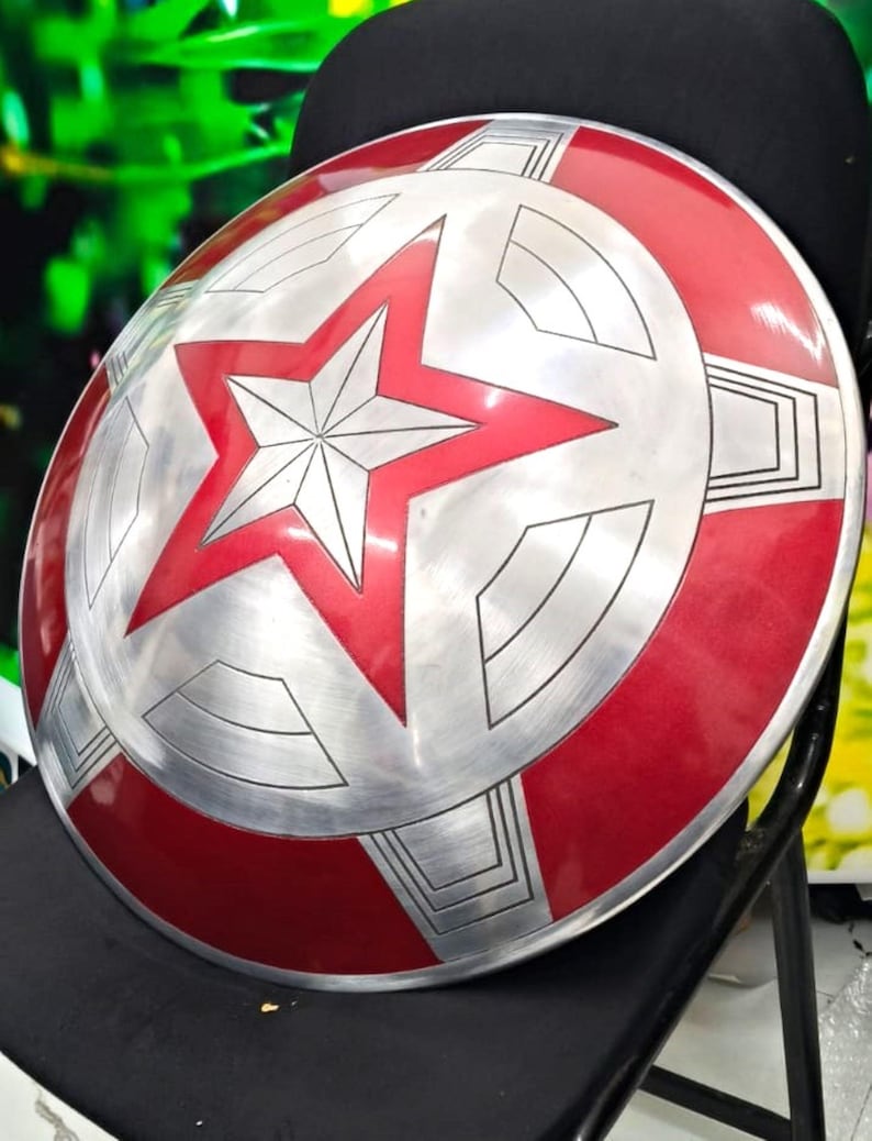 May include: A red and silver Captain America shield with a large red star in the center. The shield has a textured surface and is made of metal.
