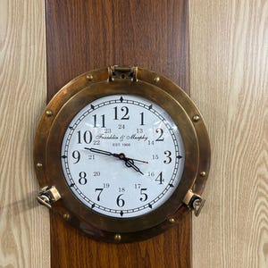 May include: A circular clock in a brass-toned frame, resembling a ship's porthole. The white clock face has black numerals and hands. The text "Franklin & Murphy EST. 1868" is visible. The clock is mounted on a wooden surface with vertical stripes.
