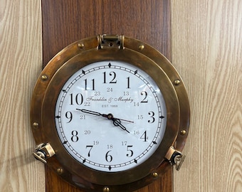 May include: A round wall clock with a brass porthole-style frame featuring a white face, black Arabic numerals from 1 to 12, and smaller inner numerals from 13 to 24. The center displays the text Franklin & Murphy EST. 1868 with black hands.