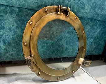 Könnte beinhalten: Runder Bullauge aus Messing mit einem transparenten Glasmittelteil. Das Bullauge hat eine aufklappbare Oberseite und einen seitlichen Riegel. Der Rahmen ist mit Nieten befestigt. Dieses Bullauge im nautischen Stil ist ein Dekorationsartikel.