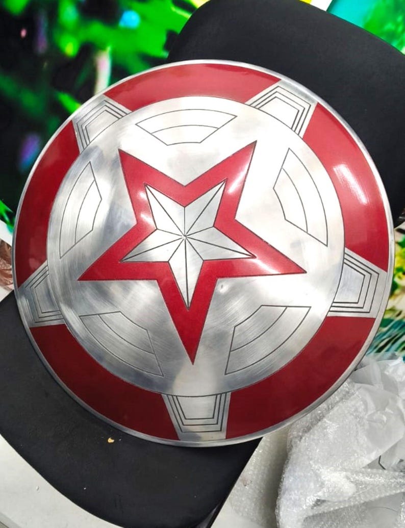May include: A red and silver Captain America shield with a five-pointed star in the center. The shield is round and has a raised edge.