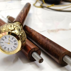 May include: A wooden walking cane with a gold-coloured clock face. The clock has Roman numerals and a white background. The cane is brown with a decorative gold-coloured handle. The cane is disassembled, showing the threaded end.