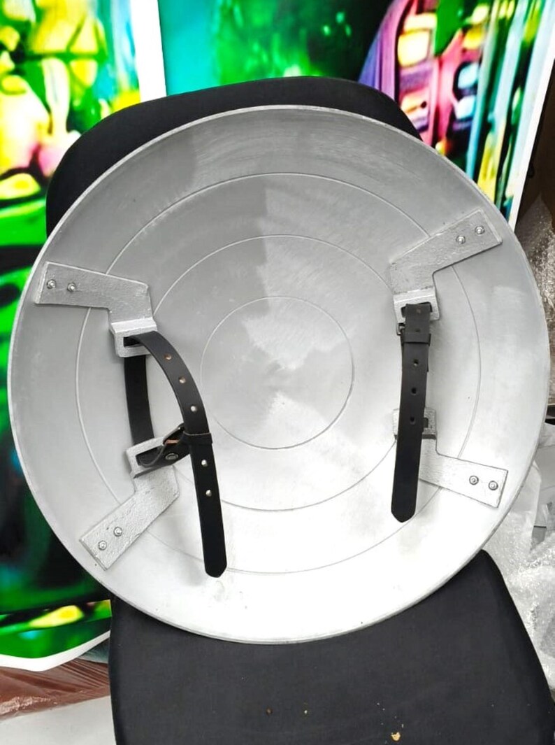 May include: The back of a silver Captain America shield with black leather straps. The shield is made of metal and has a circular design.