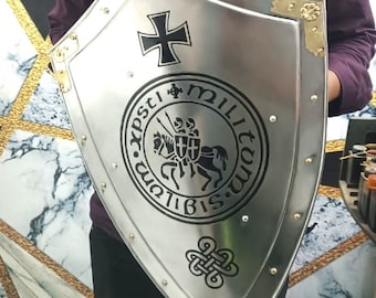 May include: A silver-colored shield with a black cross at the top and a circular emblem featuring a knight on horseback. The shield has gold-colored corner accents and is riveted around the edges.