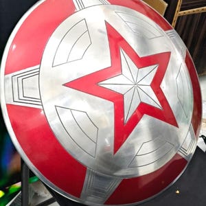 May include: A red and silver Captain America shield with a five-pointed star in the center. The shield is round and has a raised edge.