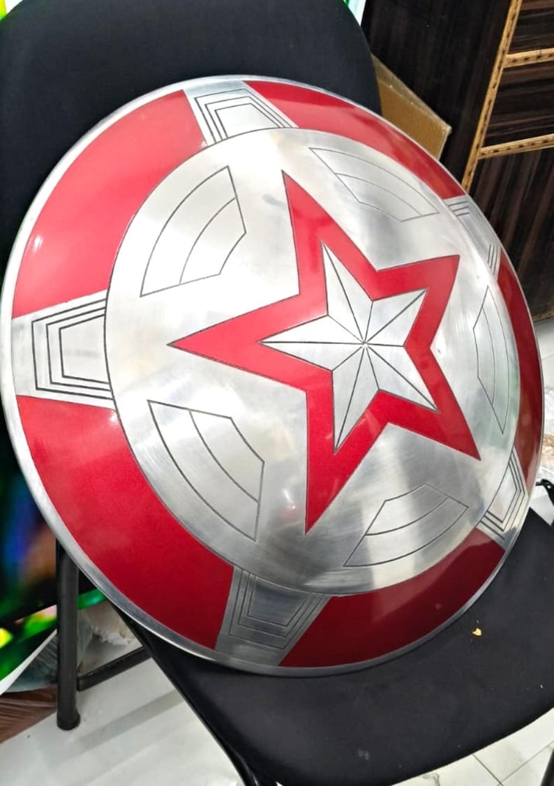 May include: A red and silver Captain America shield with a five-pointed star in the center. The shield is round and has a raised edge.