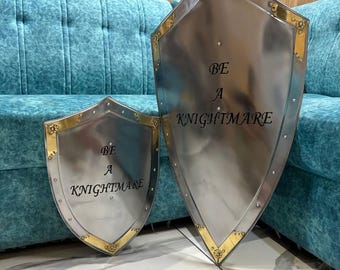 May include: Two silver-toned metal shields with gold-colored accents at the corners. Both shields feature the black text "BE A KNIGHTMARE" centered on the surface. The shields are pointed at the bottom and have visible rivets along the edges.