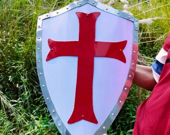 Könnte beinhalten: Ein Schild im mittelalterlichen Stil mit weißem Hintergrund und einem großen roten Kreuz in der Mitte. Der Schild hat einen silbernen Metallrand mit Nieten. Der Hintergrund ist eine Mischung aus grünem Laub und hohem, weißem Gras.