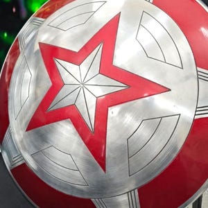 May include: A red and silver Captain America shield with a five-pointed star in the center. The shield is made of metal and has a textured surface.