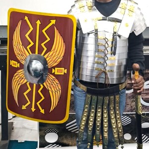 May include: A full Roman legionary armor set, including a helmet, chest plate, and shield. The shield is maroon with gold accents and a central silver boss. The armor is silver and gold, with a matching sword and belt. The outfit is a historical costume.