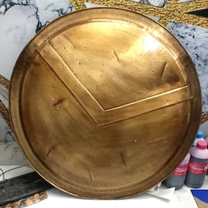 May include: A round, bronze-colored shield with a distinctive V-shaped design in the center. The shield has a raised rim and a smooth, slightly textured surface.
