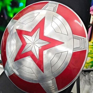 May include: A red and silver Captain America shield with a five-pointed star in the center. The shield is made of metal and has a textured surface.