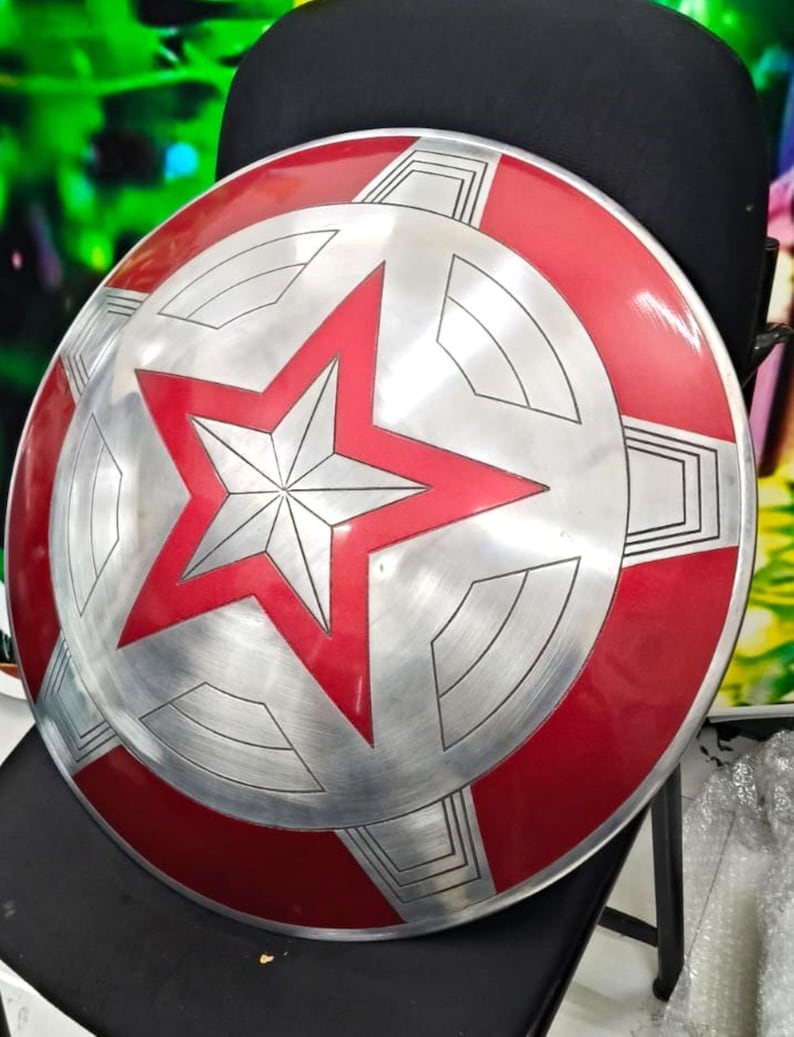May include: A red and silver Captain America shield with a five-pointed star in the center. The shield is made of metal and has a textured surface.