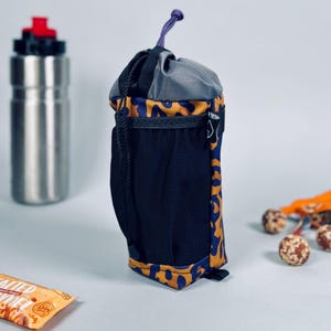 May include: A water bottle carrier with a black mesh pocket and a drawstring closure. The carrier has a pattern of orange spots on a blue background. A stainless steel water bottle with a red cap is in the background.