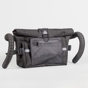 Può includere: Borsa da manubrio nera con chiusura a rotolo e tasca frontale con cerniera. La borsa è progettata per essere attaccata ai manubri della bicicletta. L'immagine mostra la borsa con i manubri attaccati e una striscia riflettente.