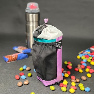 Puede incluir: Una pequeña bolsa negra con cremallera y una parte superior de cordón de color morado, turquesa y blanco. La bolsa está sobre una superficie negra con caramelos de colores esparcidos a su alrededor.