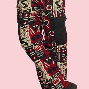 Op de afbeelding: Cargo broek met een opvallend geometrisch patroon in rood, zwart en crème. De broek heeft een zwarte zak aan de zijkant en wordt gedragen met zwarte schoenen. Het Regal Point-logo is linksonder zichtbaar.