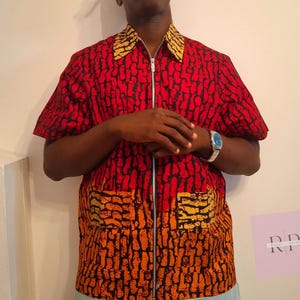 Peut inclure: Une chemise rouge à manches courtes avec un motif noir et une fermeture éclair sur toute la longueur. La chemise a un col et des poches à motifs jaunes et noirs. Une montre argentée est visible au poignet.