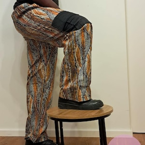 Puede incluir: Pantalones estampados con un parche negro en la rodilla. Los pantalones presentan un diseño ondulado repetitivo en naranja, marrón y plata. La persona lleva zapatos negros y una camiseta negra. Los pantalones están sobre un pequeño taburete de madera.