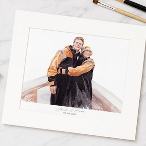 Könnte beinhalten: Aquarellporträt von zwei Personen, die sich umarmen und schwarze und orangefarbene Jacken und einen Hut tragen. Das Kunstwerk ist in einem weißen Passepartout gerahmt. Der Text "Jack & Rose" und das Datum "05.06.2025" stehen unten.