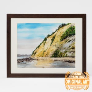 May include: Watercolor painting of a coastal cliff with a sandy beach and blue sky. The artwork is framed in a dark brown wooden frame with a white mat. The painting features yellow and green vegetation on the cliffside. The text "HAND PAINTED ORIGINAL ART NO PRINTS" is in the lower right corner.