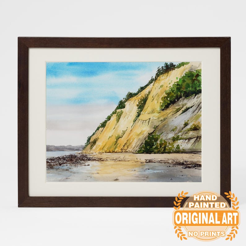May include: Watercolor painting of a coastal cliff with a sandy beach and blue sky. The artwork is framed in a dark brown wooden frame with a white mat. The painting features yellow and green vegetation on the cliffside. The text "HAND PAINTED ORIGINAL ART NO PRINTS" is in the lower right corner.