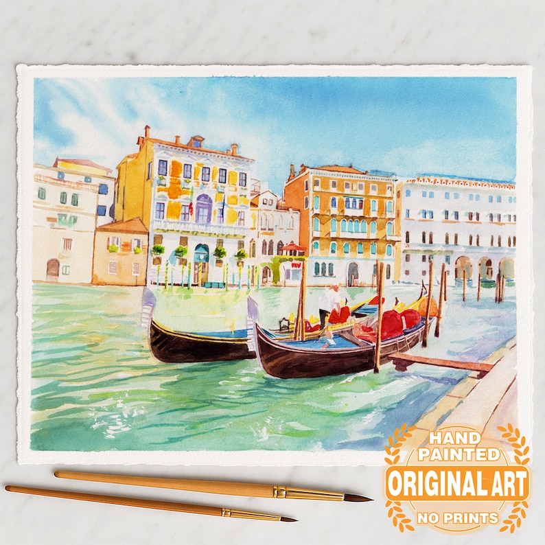May include: Watercolor painting of Venice, Italy, featuring gondolas on a canal with colorful buildings in the background. The artwork is hand-painted and labeled as original art, with no prints. Two paintbrushes are in the foreground.