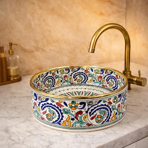 May include: A round ceramic basin with a vibrant floral and paisley pattern. The basin has a gold rim and sits on a marble surface. A gold tap is positioned above the basin.