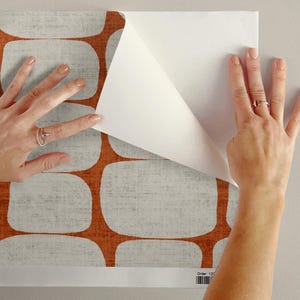 May include: A sheet of peel-and-stick wallpaper featuring a repeating pattern of rounded rectangular shapes in a textured light gray color separated by thick, solid burnt orange lines. The paper has a white adhesive backing partially peeled back.