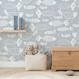 May include: A light blue wall with a repeating white fish pattern. A wooden shelf holds books. A canvas basket with wooden handles contains stuffed animals. A wooden chest sits on the right. A round rug is on the floor.