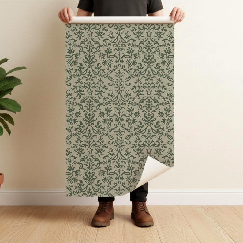 A person is standing in front of a wall, holding up a roll of wallpaper with a repeating green and white floral pattern.