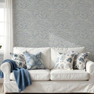 May include: A white sofa featuring four decorative throw pillows with blue and white botanical and wave patterns, accompanied by a solid blue knit throw blanket. The background wall is covered in wallpaper with a repeating blue wave pattern.