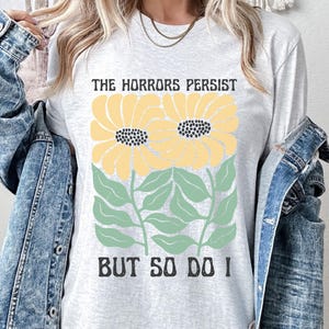May include: A gray long-sleeved t-shirt with a graphic of two yellow flowers with green leaves and the text "The Horrors Persist But So Do I".