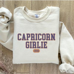 May include: A cream-colored sweatshirt with the text "Capricorn Girlie 1990" in navy blue and orange lettering.