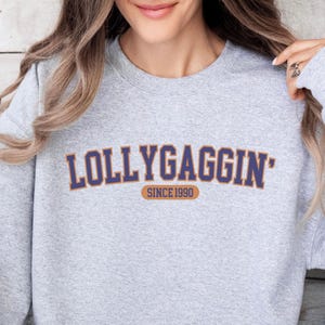 May include: Gray sweatshirt with the text "LOLLYGAGGIN' SINCE 1990" in a navy blue and orange collegiate font.