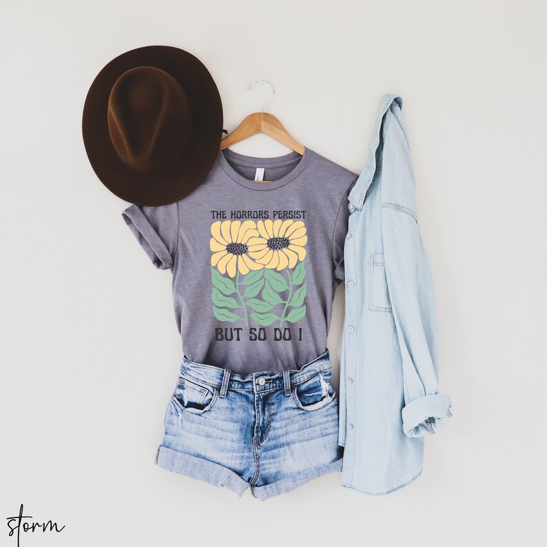 May include: A gray t-shirt with a graphic of two yellow sunflowers with green leaves and the text "The horrors persist but so do I".