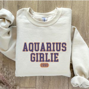 May include: A cream colored sweatshirt with the text "Aquarius Girlie 1990" in blue and orange lettering.