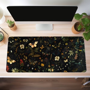 May include: A rectangular desk mat featuring a dark background with a botanical design. The design includes butterflies, flowers, and foliage in shades of yellow, orange, and green. The mat is placed on a light-colored wooden desk.