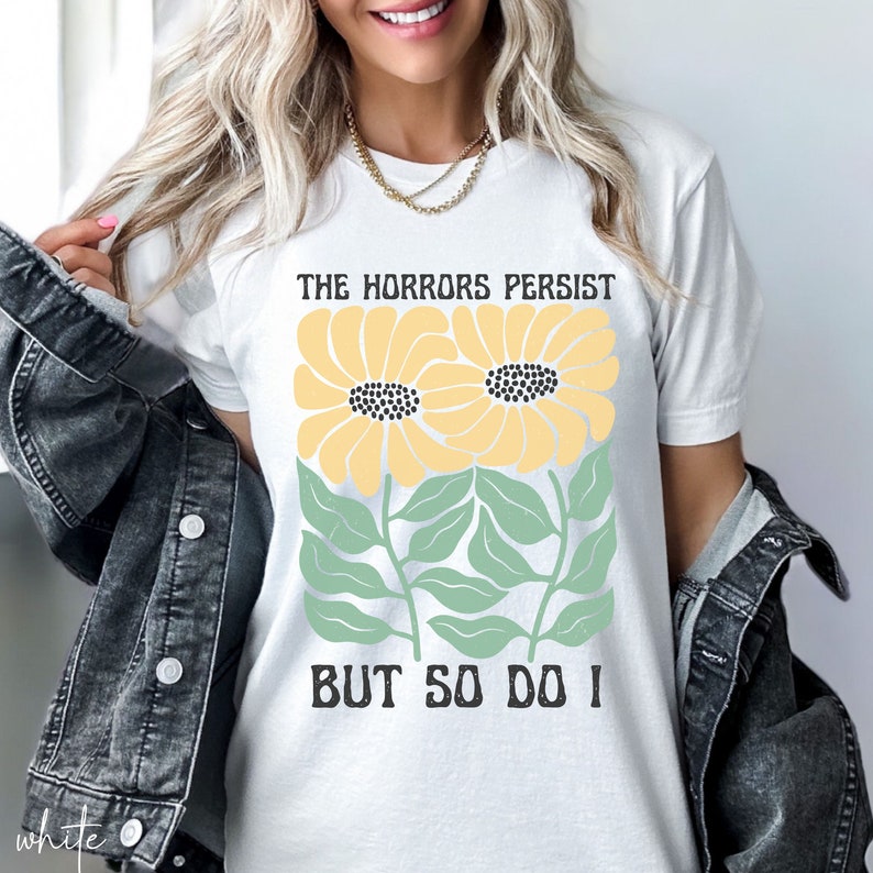 May include: White t-shirt with a graphic of two yellow sunflowers with green stems and leaves. The text "The Horrors Persist But So Do I" is printed on the shirt.