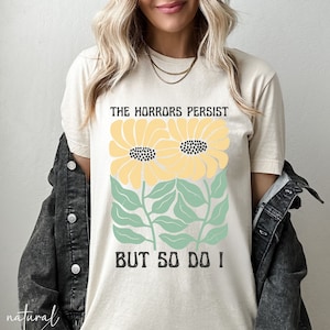 May include: A beige t-shirt with a graphic print of two yellow sunflowers with green leaves and the text "The Horrors Persist But So Do I".