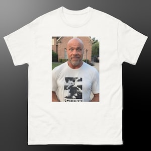 Puede incluir: Camiseta blanca con una imagen en blanco y negro de una persona y la palabra "SCHULTZ" en un recuadro rectangular. La persona de la imagen lleva la camiseta. La camiseta está hecha de un material suave y cómodo.