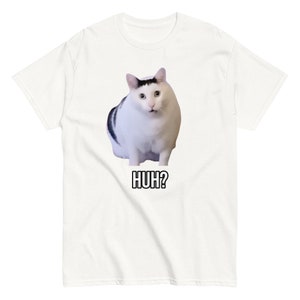 May include: White t-shirt featuring a meme of a cat with black markings and the text "HUH?" in a black font. The cat has a surprised expression. The t-shirt is a short-sleeved, crew neck style.