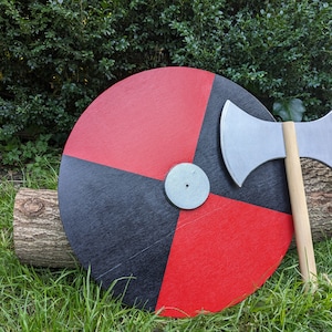 May include: A black and red round shield with a silver axe resting on top of it. The shield is on a bed of green grass.