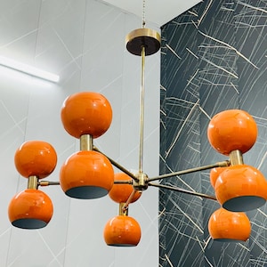 May include: A mid-century modern chandelier with eight orange globe shades. The fixture has a brass finish and hangs from a chain. The shades are a glossy orange and are evenly spaced around the central hub.