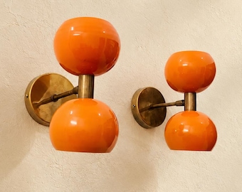 May include: Two wall-mounted light fixtures featuring a brass circular base and arm, each supporting two glossy orange spherical glass shades stacked vertically. The design includes a central brass rod connecting the two orange globes on each unit.