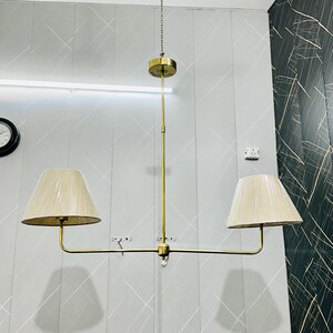 May include: A brass-colored, two-light chandelier with beige lampshades. The fixture hangs from a chain and features a central stem with two arms extending to hold the lampshades. The lampshades are conical and made of a textured fabric.