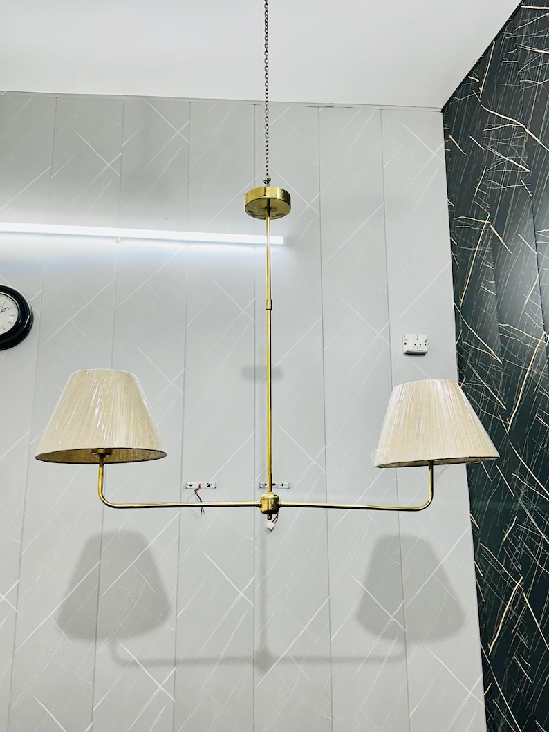 May include: A brass-colored, two-light chandelier with beige lampshades. The fixture hangs from a chain and features a central stem with two arms extending to hold the lampshades. The lampshades are conical and made of a textured fabric.